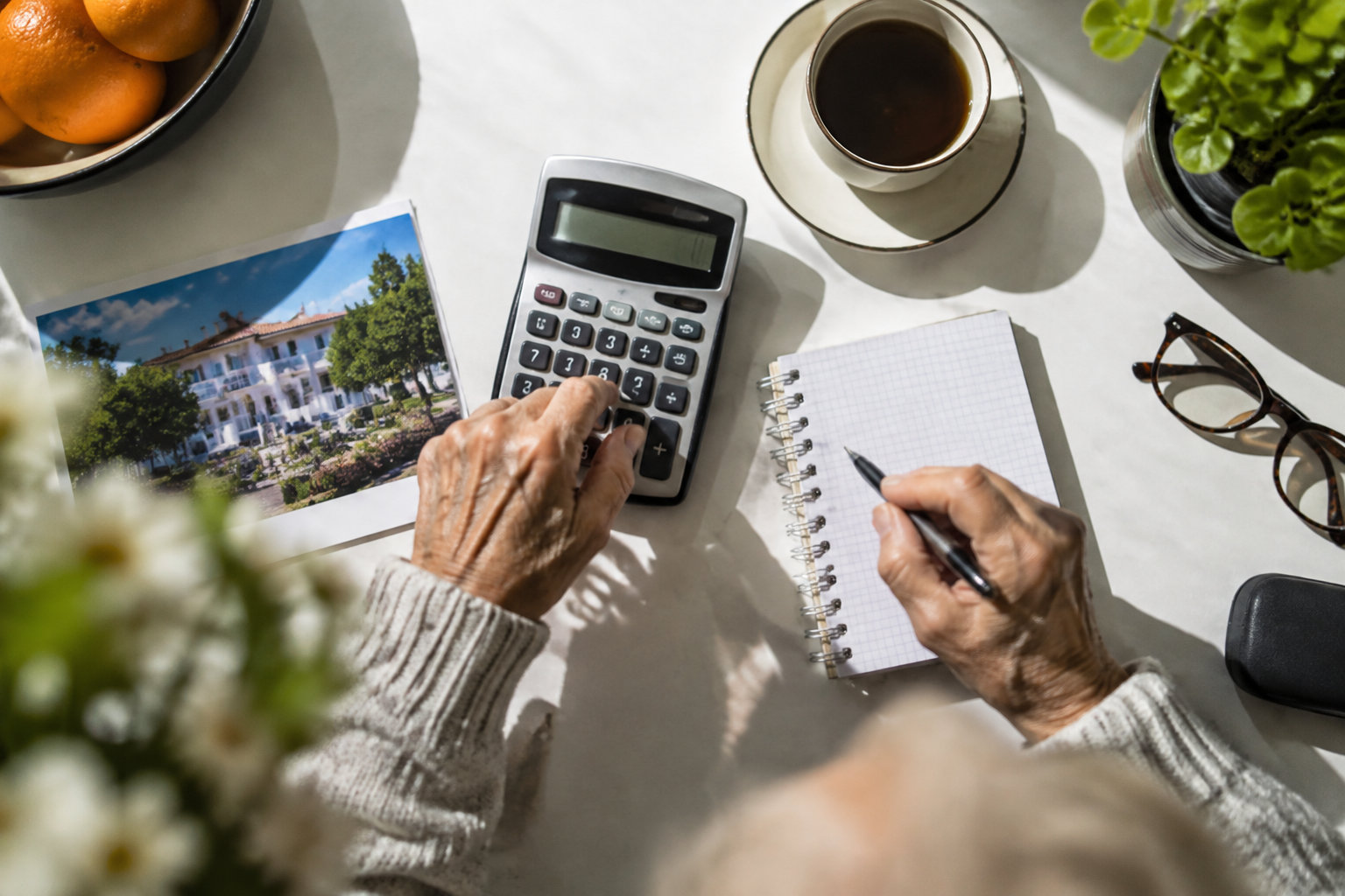Budgétiser une maison de retraite au Portugal Vue de dessus d'une personne âgée utilisant une calculatrice et un carnet à côté d'une photo de villa portugaise.